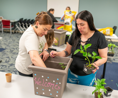 Two Native American women place plants in a square pot with dirt that says "welcome" - the pot is on a table in a carpeted classroom