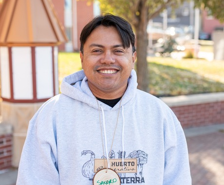 Latino man looking at the camera, wearing a white hooded sweatshirt, a smile, and a wooden name tag that says "Sandro"