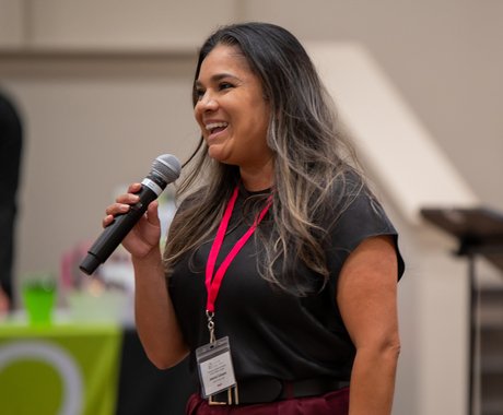 Woman with long brown hair, wearing a black short sleeved shirt, holding a microphone, smiling toward the left of the photo