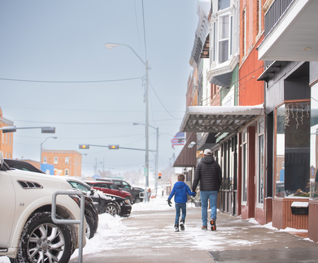 A dad and son hold hands while walking down a small town main street sidewalk. They're wearing coats and snow is swirling on the ground. 