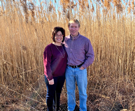 Man and woman standing in front of tall grass in a field. Both are smiling at the camera.