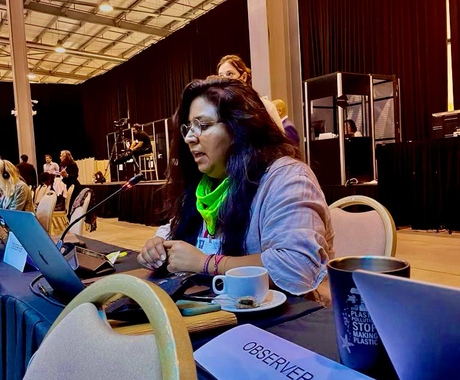 A person with long brownish-black hair, wearing bright green buff and a quarter sleeve button up shirt, speaks into a microphone while seated at a table. In front of them is a computer, a coffee cup, and a name tag that says 'observer' 