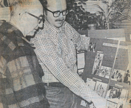 Foto de diari antiga (de la dècada de 1970) de dos homes. Un home amb un abric a quadres està mirant un altre home amb una camisa de màniga llarga amb botons a quadres assenyalant un cartell amb fotos.