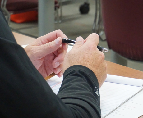 Les mans d'una persona portant mangas llargues de color negre, detienen un bolígrafo a sobre d'un cuaderno blanc en una taula cafè