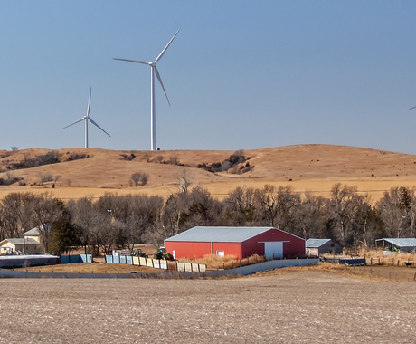 A farm seen across a harvested field, with a red machine shed surrounded by a fence plus a house and several outbuildings. Four wind turbines are behind the farm on a brown hillside.