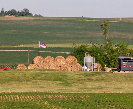 a US flag standing tall over a pile of round hay bales, stacked 2 high. In the foreground and background, there are green fields with short crops.