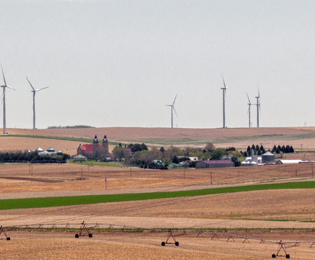 a distant view of a small town in the middle of brown harvested fields, with wind turbines dotting the hills above and behind the town