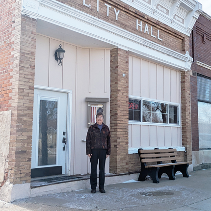 Woman with short dark hair, glasses, a dark jacket, and black pants smiles outside of a brick building labeled "CITY HALL."