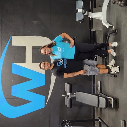 A hispanic male wearing a black shirt and grey shorts stands next to a hispanic female wearing a blue shirt and black pants inside a gym with gym equipment around them