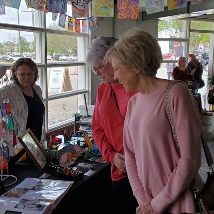 Una mujer está parada detrás de un stand con obras de arte mientras dos mujeres mayores con camisas rosas de manga larga miran por encima de la mesa.