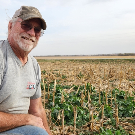 Man kneeling in field, with green plants growing among corn stalks. Man is wearing a gray Tshirt, ball cap, sunglasses, and has a white beard and mustache.