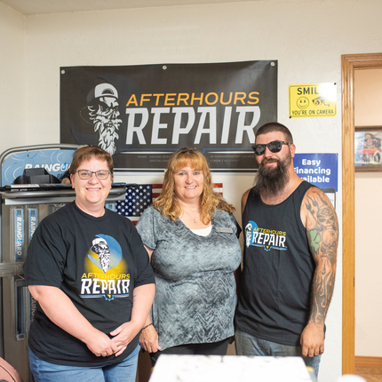 Tres persones: una dona a la dreta i un home a l'esquerra amb samarretes negres "Afterhours Repair" i una dona al mig amb una camisa grisa de màniga curta, dempeus en una oficina davant d'un cartell que diu "Afterhours Repair".