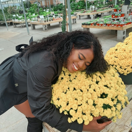 Una dona afroamericana que porta una jaqueta de màniga llarga i unes botes negres altes abraça un test de flors grogues en un hivernacle