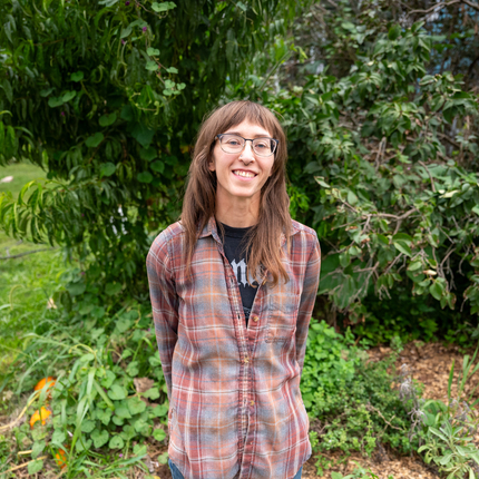 Woman standing in front of a tree/bush. Woman has brown hair with bangs, glasses, a reddish plaid shirt and is smiling directly at the camera.