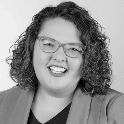 Mujer sonriendo a la cámara, con cabello castaño rizado hasta los hombros, gafas y una chaqueta sobre una camisa oscura.