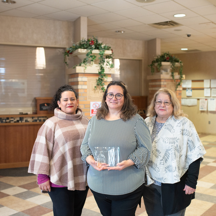 Tres mujeres posan para una foto en el vestíbulo de una oficina con un galardón en mano।
