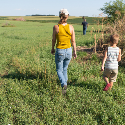 White woman with a mustard yellow tanktop and light blue jeans and a baseball cap walks along side a child wearing a stripped white and grey tanktop and beige shorts in a large field