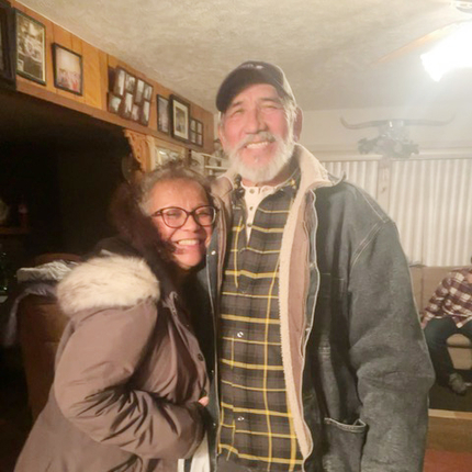 Un hombre y una mujer latinos en edad de jubilación sonriendo cálidamente a la cámara desde el interior de su casa, vestidos para el clima frío.