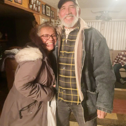 Un hombre y una mujer latinos en edad de jubilación sonriendo cálidamente a la cámara desde el interior de su casa, vestidos para el clima frío.