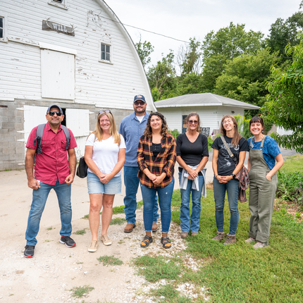 A group of 7 people standing in front of a white barn. The people range in age and gender and are wearing casual farm-visiting clothes like jeans and T-shirts.