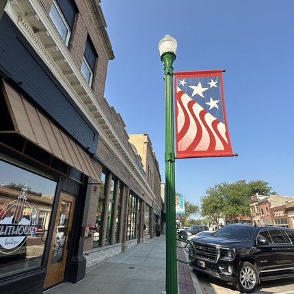 Main street with businesses on the left, a light pole with a flag banner and cars parked along the street