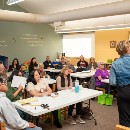 Group of more than 10 men and women sit at long tables facing towards a white woman with short blond hair wearing a jean jacket and black dress presenting to the group in a medium size room with green and yellow walls