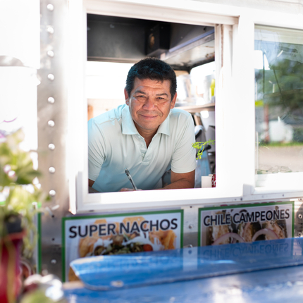 Hombre hispano de mediana edad con cabello negro corto, asomado a la ventana de un camión de comida, esperando tomar un pedido y sonriendo a la cámara.