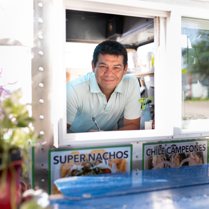 Hombre hispano de mediana edad con cabello negro corto, asomado a la ventana de un camión de comida, esperando tomar un pedido y sonriendo a la cámara.