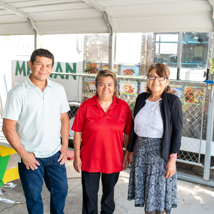 Tres personas de mediana edad, un hombre y dos mujeres, de pie frente a su camión de comida y al área cubierta para comer, sonriendo a la cámara.