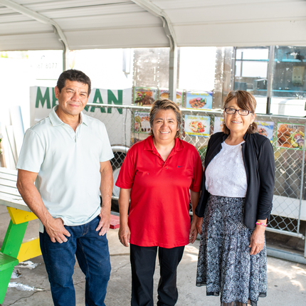 Tres personas de mediana edad, un hombre y dos mujeres, de pie frente a su camión de comida y al área cubierta para comer, sonriendo a la cámara.