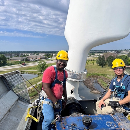 Two men wearing yellow hard hats sit on top of a wind turbine