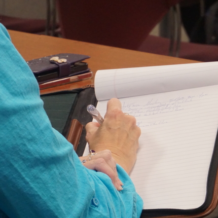 A white individuals right arm with a blue sleeve, and their hand writing on a white notepad near a wooden table