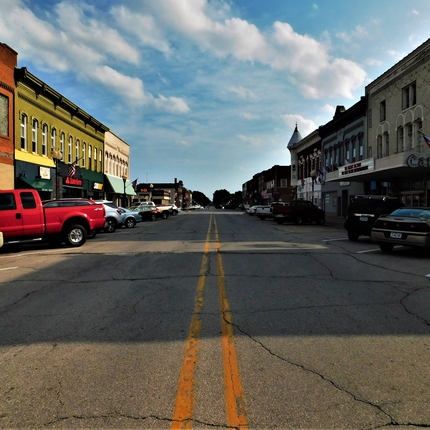 View of a main street in rural community, with buildings along the left and right side, vehicles parked in front of storefronts, cracked concrete street