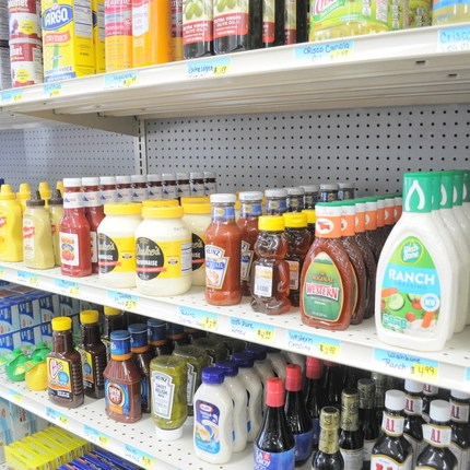 Estantes de comida en una tienda de comestibles de un pueblo pequeño. En primer plano, salsa ranch, ketchup, mayonesa, miel y, en el extremo izquierdo, papel de aluminio y bolsas de plástico.