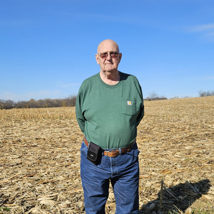 Man standing in a field covered in corn stalks. Man is wearing a green long-sleeved shirt, blue jeans, sunglasses, and has no hair.