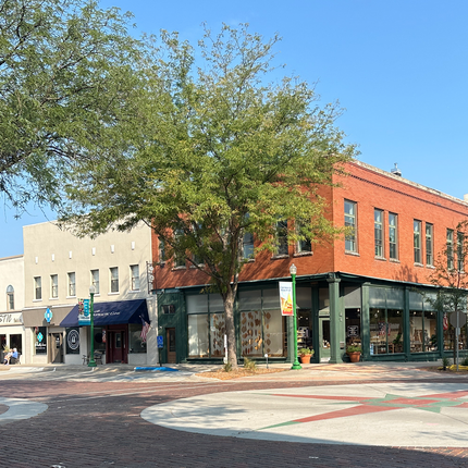 Main street corner in Ashland, NE. There is a tall, two-story red brick building with green posts on the corner with a two-story white brick building next to it. The red brick road has a cement circle at the intersection, with a red and green mosaic image in the center. 