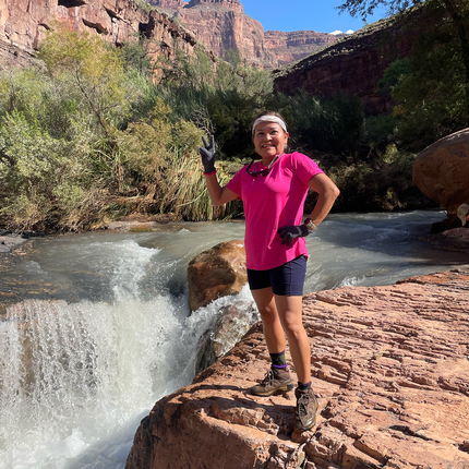 Indigenous woman wearing a pink shirt, black shorts, and black gloves, gives a peace sign while standing on red rock at the lip of a waterfall