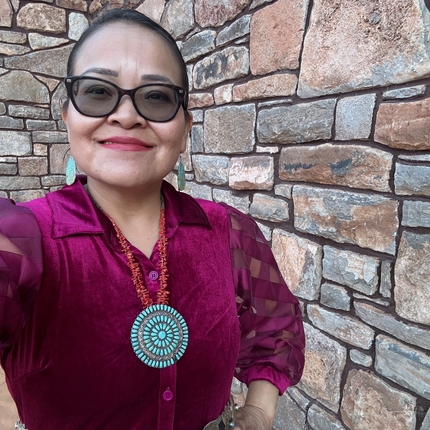 Indigenous woman looking at the camera, wearing sunglasses and a beaded necklace, as well as a purple dress, standing in front of a brick wall