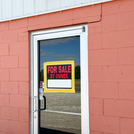 Edifici de maó de color salmó amb una porta comercial clara al mig. Un cartell "en venda per propietari" a la part superior central de la porta.