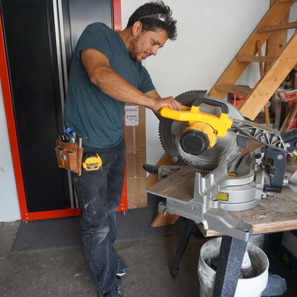 Un home hispà que porta una camisa de màniga curta de la marina, pantalons negres i un cinturó d'eines al voltant de la cintura utilitza una gran eina de tall per tallar un panell de parquet dins d'un garatge