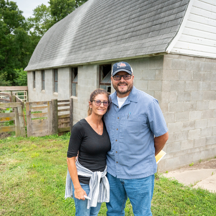 Man and woman standing in front of an old gray brick barn. Woman is wearing a black quarter sleeve shirt, glasses, and a light sweater tied around her waist. Man is wearing glasses, baseball cap, and a blue plaid button up shirt. 