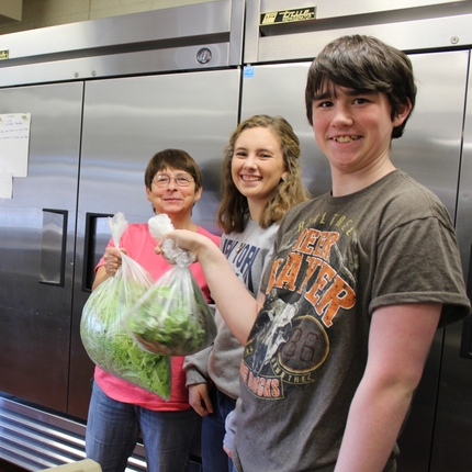 A lunch lady in a pink shirt holds a big bag of freshly cut lettuce, accompanied by two high school students - a boy and a girl - standing in front of industrial sized refrigerators in a school kitchen