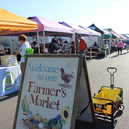 A farmers market with a row of colorful tents with tables and people underneath. In the foreground, a sign that says "Welcome to our Farmers Market" and an empty yellow wagon.