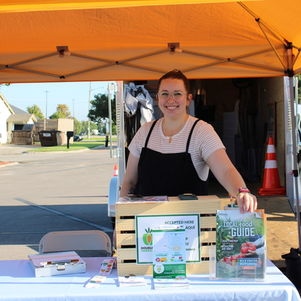 Woman in her 20s standing behind a table and display with local food resources. She's under a yellow tent at a farmers market.