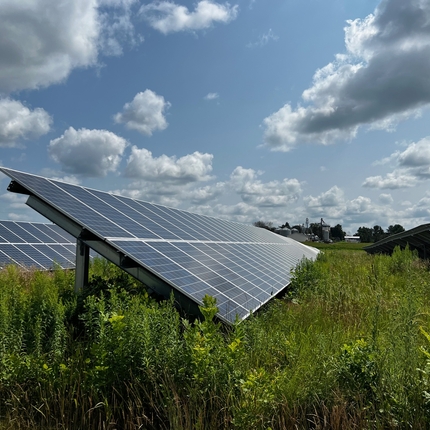 Two long solar arrays facing toward the right of the camera, with crops underneath. Sky is blue with fluffy clouds.