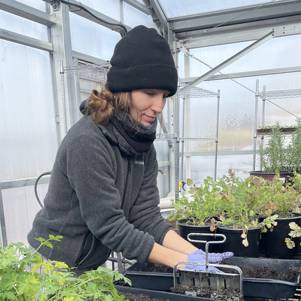 Woman in a gray fleece and black stocking cap has her hands in soil in the middle of a greenhouse. On either side of her are green plants.