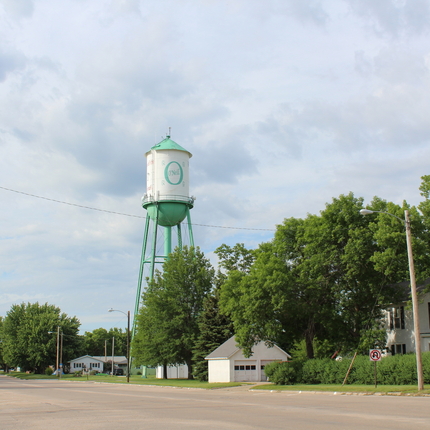 A road leading into a community, with a white house and green and white water tower on the right.