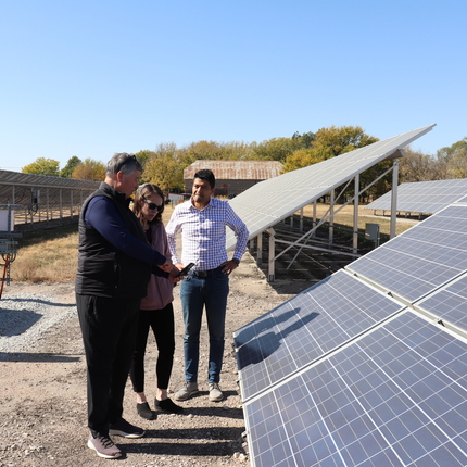 Three individuals, a woman and two men stand near solar panels while looking at a cell phone.