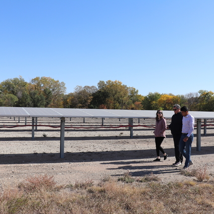 Three people walking in front of several rows of solar arrays. Green and orange trees in the background.