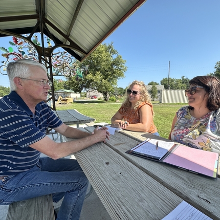 Man with gray hair wearing a blue polo with white stripes seated at a table talking with two women